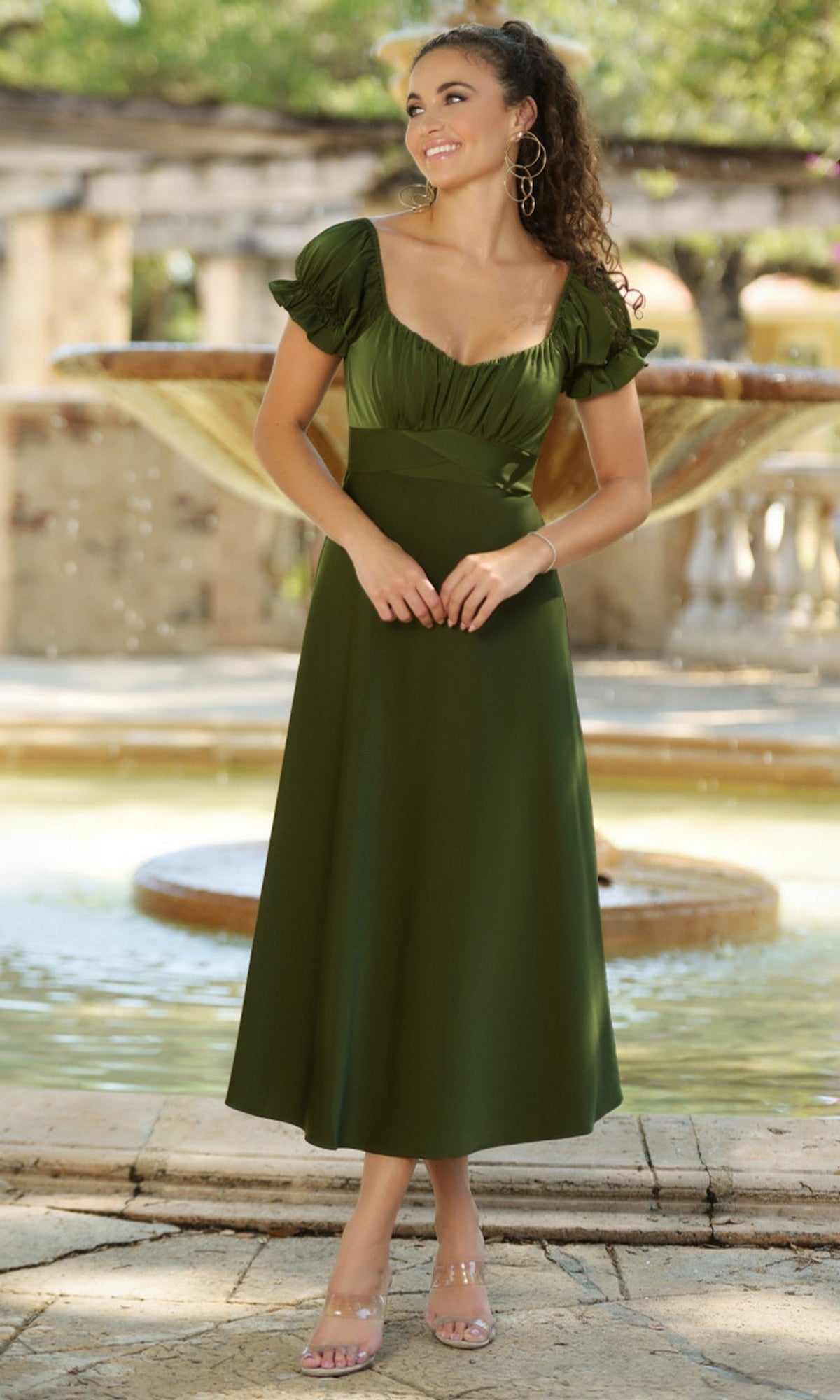 A woman wearing the Velvi Tea-Length Wedding-Guest Dress Maldives in dark green stands by an outdoor fountain, smiling and looking to the side.